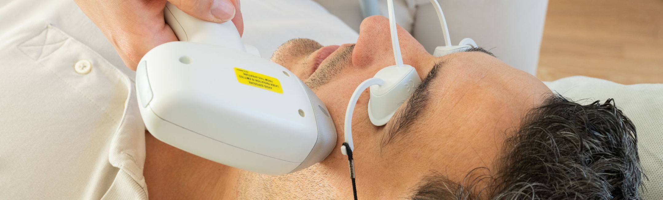 Ein Mann liegt entspannt auf einer Behandlungsliege und erhält eine kosmetische Laserbehandlung im Gesicht. Er trägt eine Schutzbrille, um die Augen vor dem Licht zu schützen, während die Therapeutin das Lasergerät professionell und sorgfältig an seiner Wange positioniert.