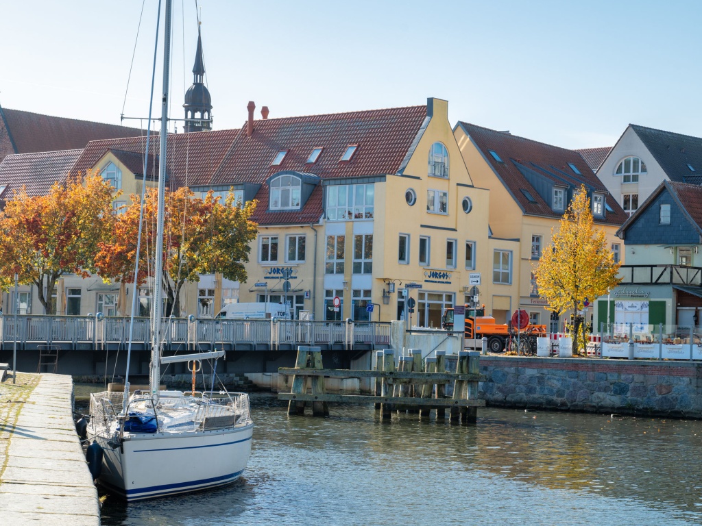 Blick auf eine Uferpromenade in Stralsund mit herbstlich gefärbten Bäumen und einem kleinen Segelboot, das am Kai festgemacht ist. Im gelben Gebäude im Hintergrund befindet sich das Complenetarium, die Heilpraxis für Osteopathie und Lasertherapie.