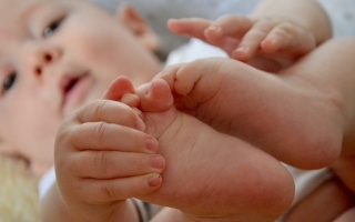 Ein Baby liegt auf dem Rücken und hält mit seinen Händen die eigenen Füße. Das Baby schaut neugierig Die Szene wirkt verspielt und zeigt die natürliche Beweglichkeit und Neugier des Babys – ein Hauptaugenmerk in der Osteopathie: Beweglichkeit und Körperwahrnehmung.